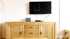 A room with a wooden table, a TV mounted on the wall, a cabinet with drawers, and a floor lamp.