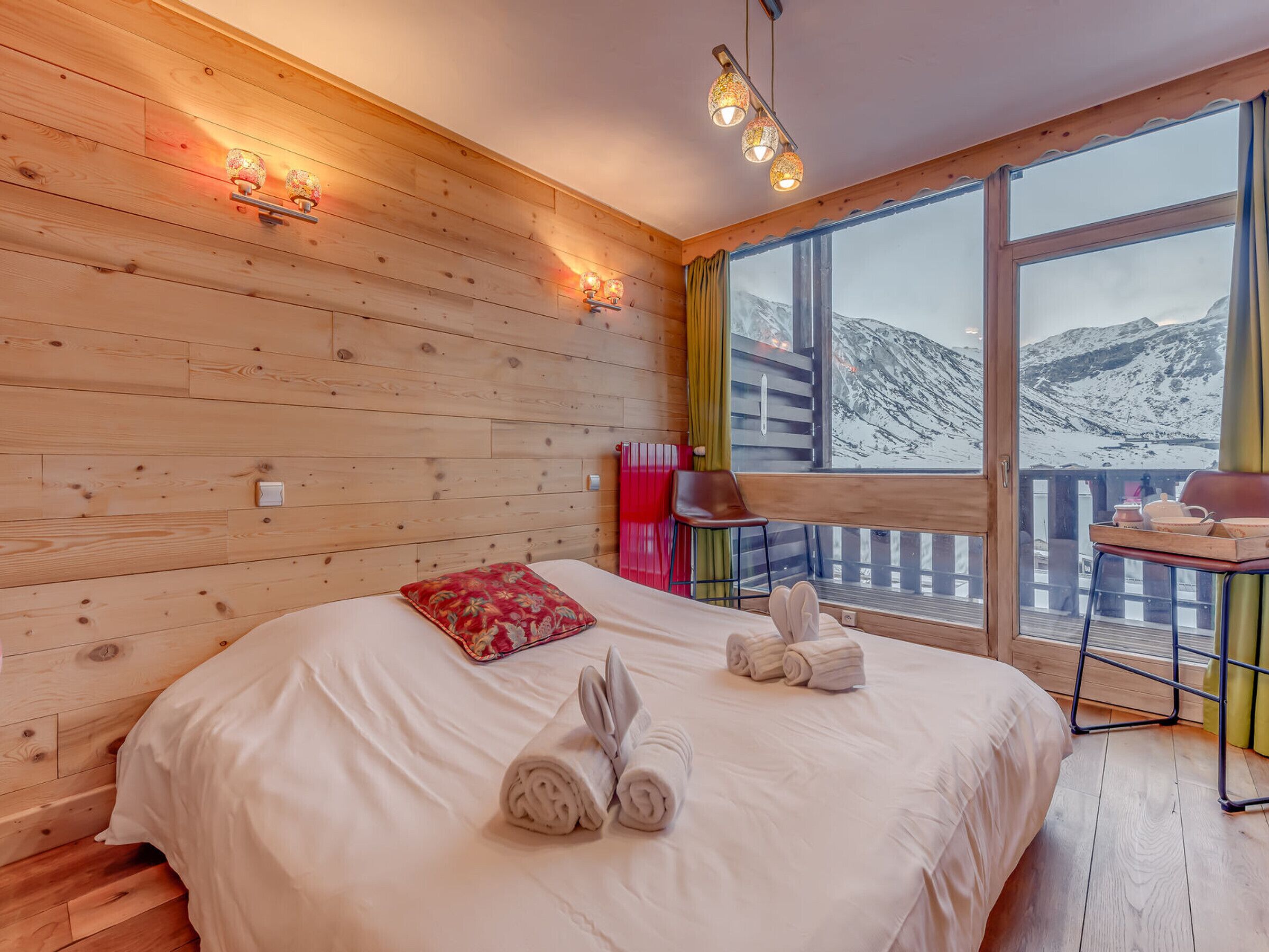 Une chambre à coucher aux murs lambrissés, avec un grand lit, un oreiller rouge et des serviettes sur le lit. Il y a une fenêtre donnant sur les montagnes et un balcon avec une table et des chaises.