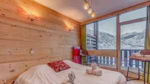 Une chambre à coucher aux murs lambrissés, avec un grand lit, un oreiller rouge et des serviettes sur le lit. Il y a une fenêtre donnant sur les montagnes et un balcon avec une table et des chaises.