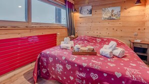 Une pièce chaleureuse aux murs en bois, avec un grand lit, une tête de lit rouge et une fenêtre donnant sur un paysage enneigé.
