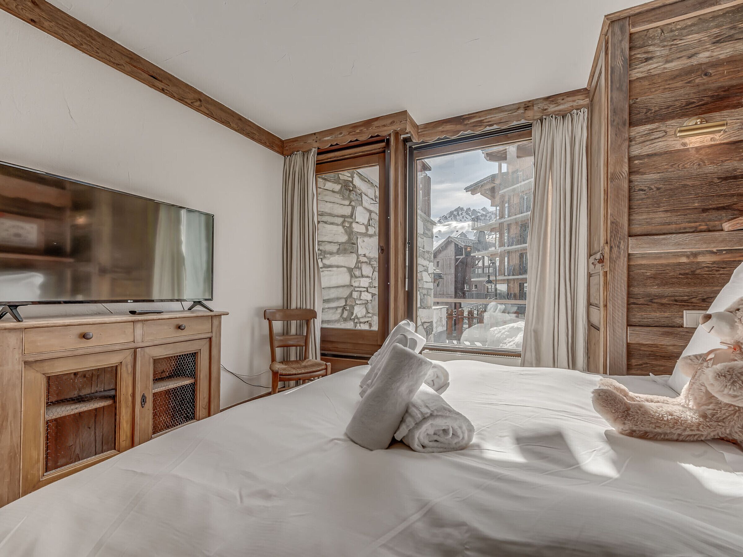 Habitación de hotel con cama, cómoda de madera, televisor y vista a un pueblo cubierto de nieve.