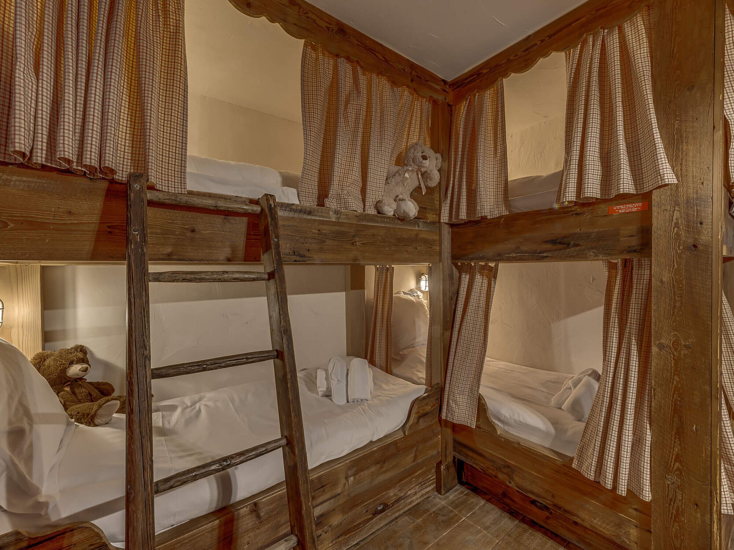 Une chambre avec des lits superposés, des cadres en bois, des rideaux à carreaux et un ours en peluche sur le lit du haut.