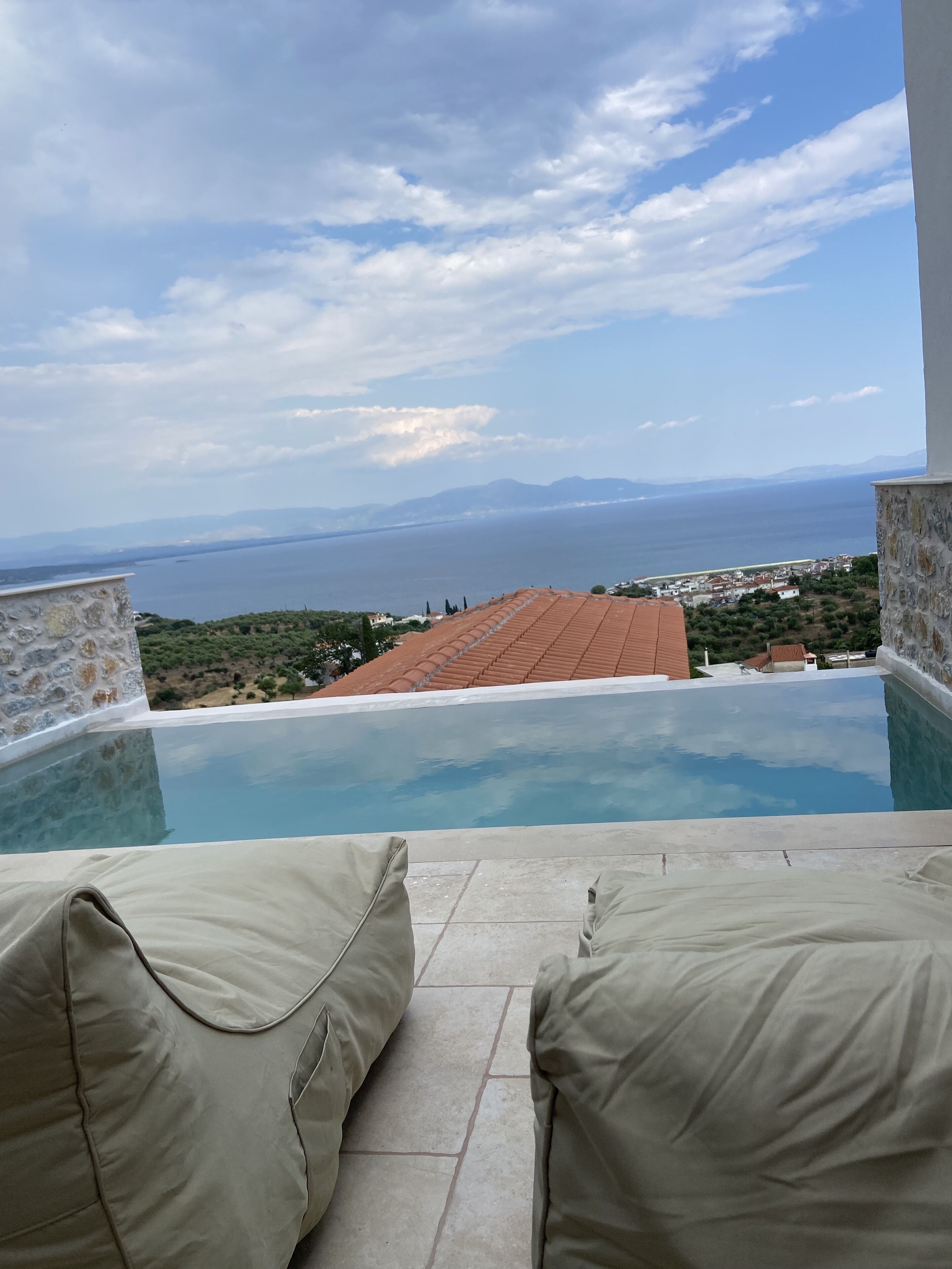 A pool with a view of the sea and mountains, surrounded by stone walls and beige cushions.