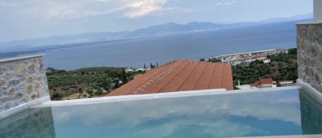 A pool with a view of the sea and mountains, surrounded by stone walls and beige cushions.