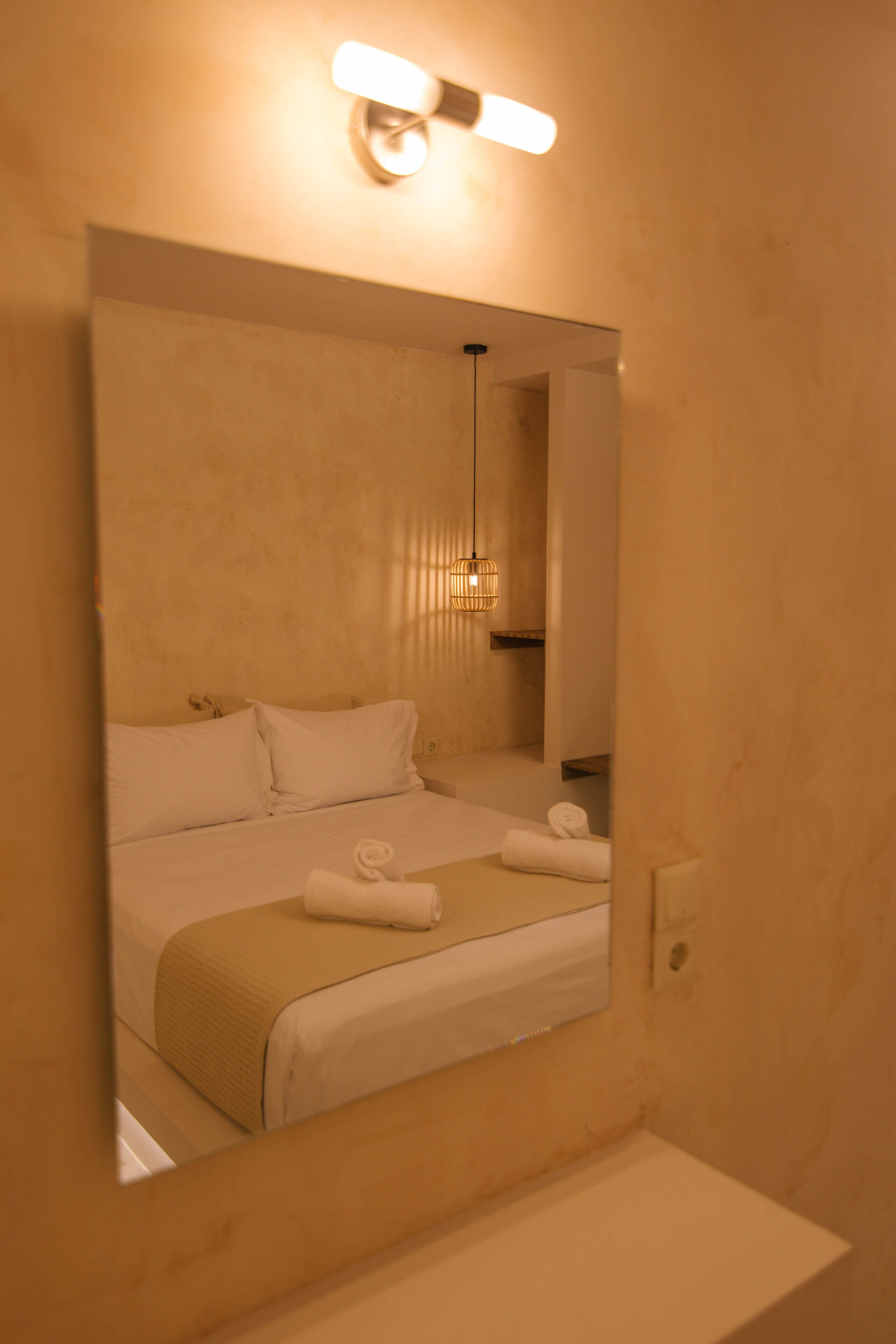 Une chambre d’hôtel dotée d’un grand miroir qui reflète un lit recouvert de draps blancs, une table de chevet et un luminaire suspendu.