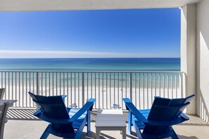 A balcony with two blue chairs and a small table, overlooking a beach with clear blue water and a sandy shore.
