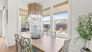 A dining area with a wooden table, white chairs, a hanging light fixture, and a potted plant. - Dancing Heron - West End PCB, Pool + Golf Cart 🛺, Sleeps 12 (Panama City Beach)