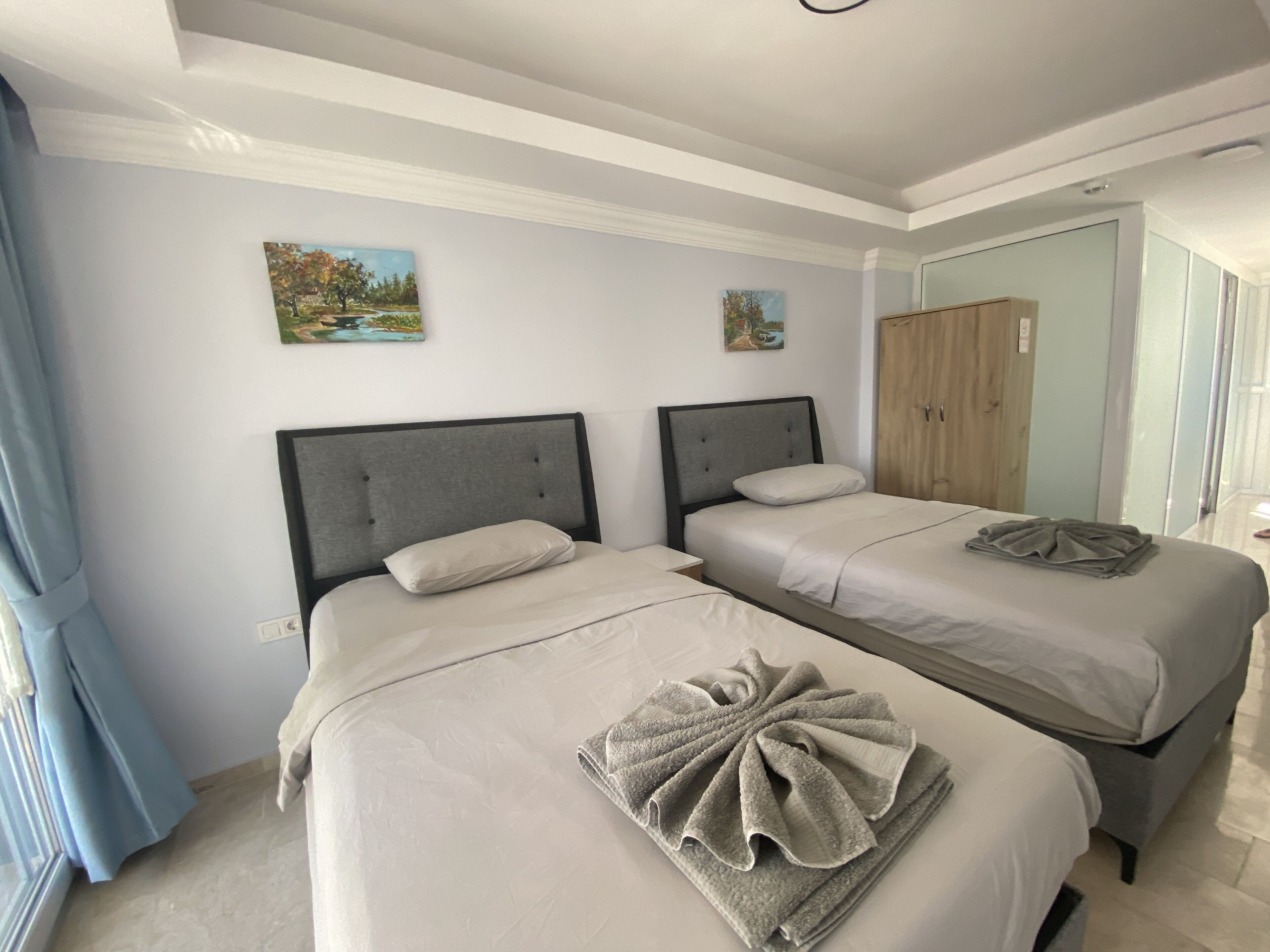 Een hotelkamer met twee bedden, elk met een grijze hoofdbord en witte beddengoed. Er hangen twee schilderijen aan de muur, een houten garderobe en een badkamer is zichtbaar door een open deur.