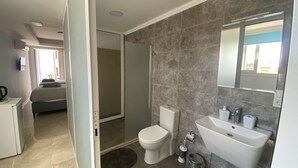Un baño moderno con inodoro, lavamanos y ducha. Hay un bote de basura pequeño y una botella de desinfectante para manos. El baño tiene suelo de baldosas y un espejo sobre el lavamanos.
