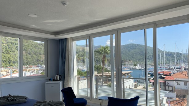 Habitación de hotel con una cama, dos sillas, una mesita y vistas a un puerto deportivo y colinas.