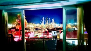 Vista desde el interior de un edificio, a través de amplios ventanales, hacia una escena nocturna con un puente y diversas luces.
