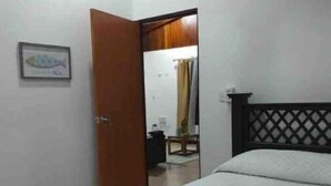 Quarto com estrutura de cama em madeira, colchão branco e cabeceira de madeira. Há uma porta que dá para um banheiro com espelho e toalha pendurada na parede.
