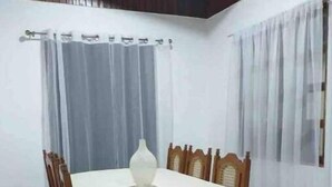Uma área de refeições com uma mesa redonda coberta por toalha branca, cercada por cadeiras de madeira.