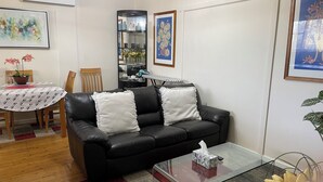 Ein Wohnzimmer mit einer schwarzen Ledersofa, zwei weißen Kissen, einem Glastisch, einem Esstisch mit Stühlen und einem Bücherregal.