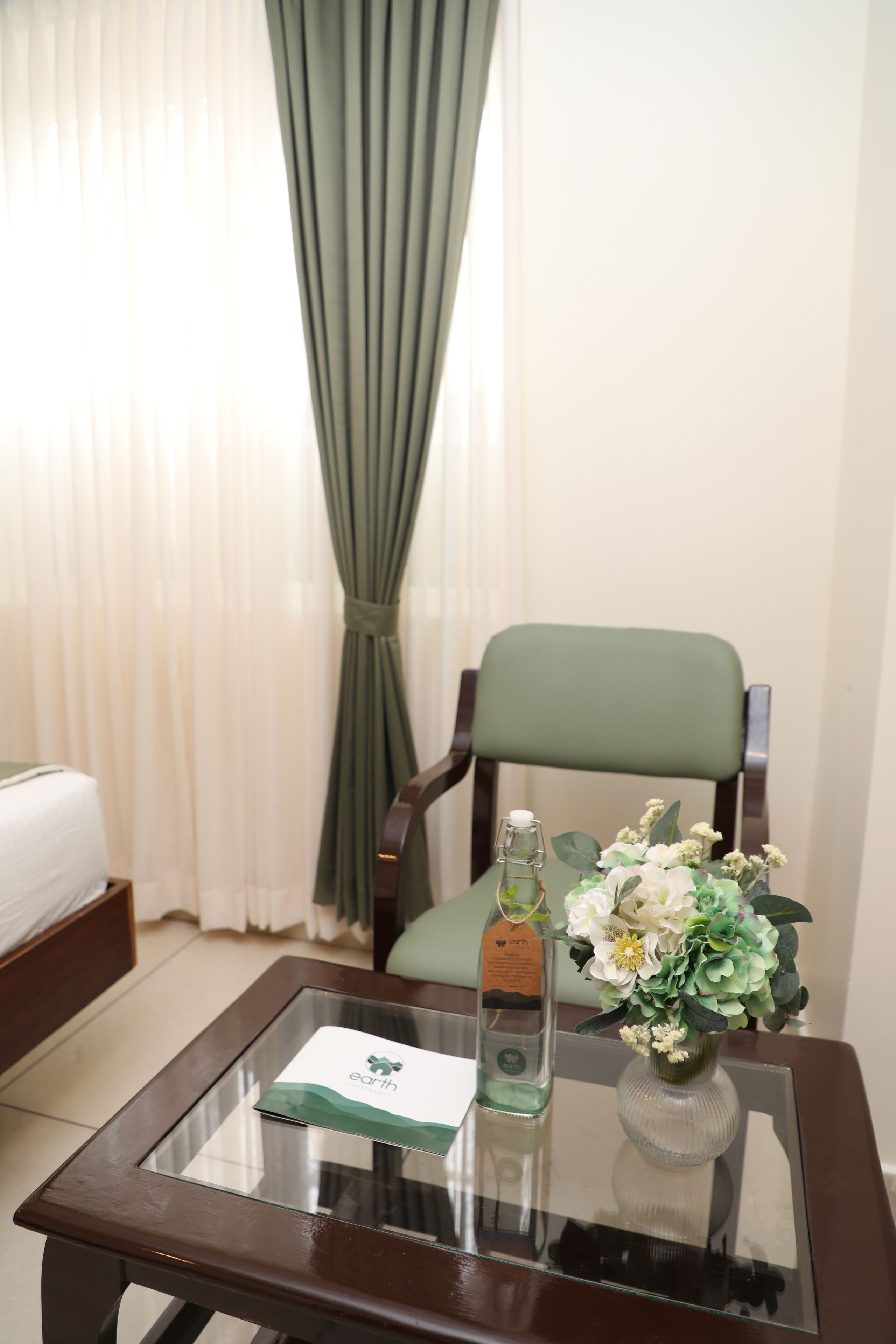 A hotel room with a bed, a green upholstered chair, a glass-top table with a bottle and a vase of flowers, and a card on the table.