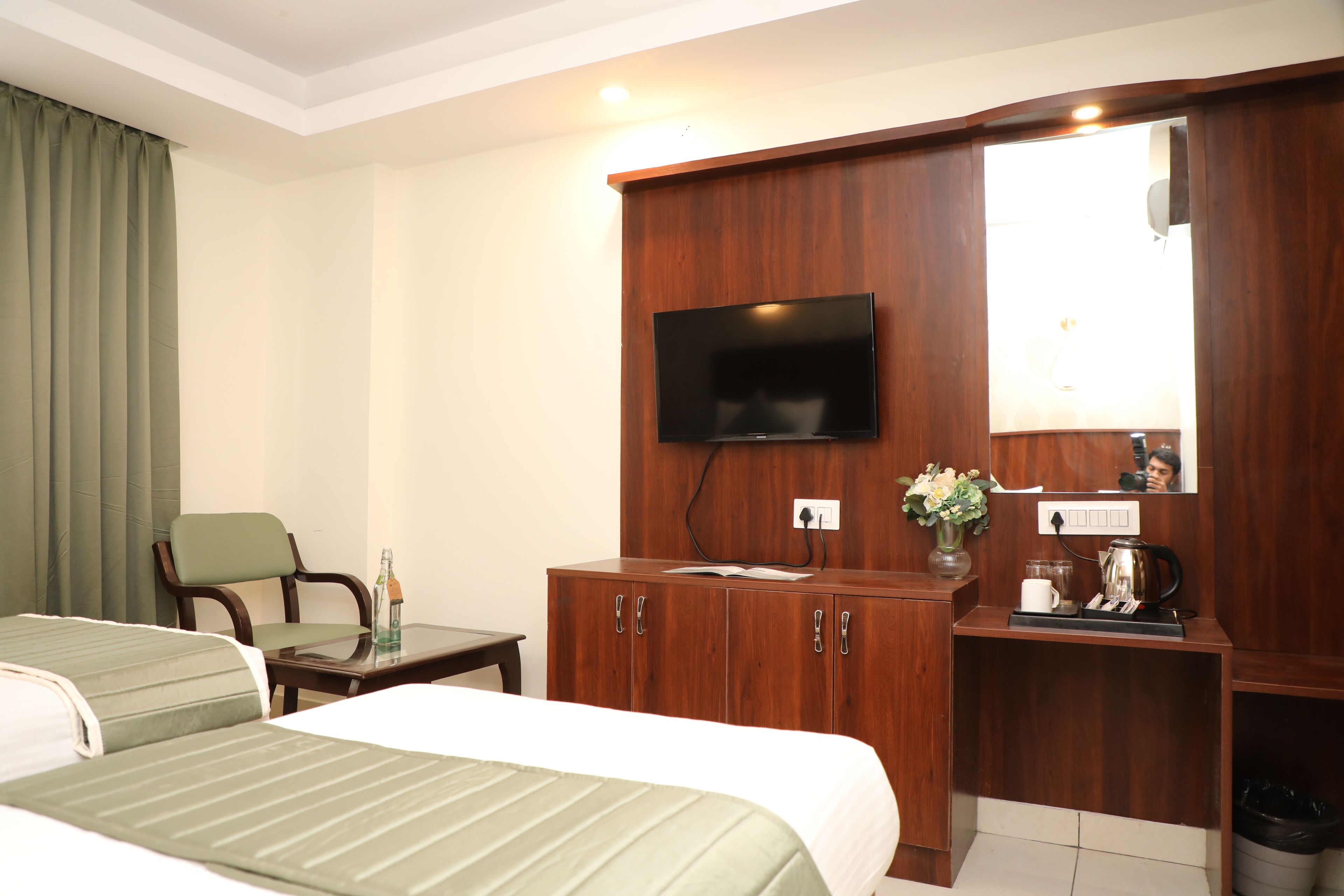 A hotel room with two beds, a TV mounted on a wooden cabinet, a small table with a bottle and papers, and a mirror above the cabinet.