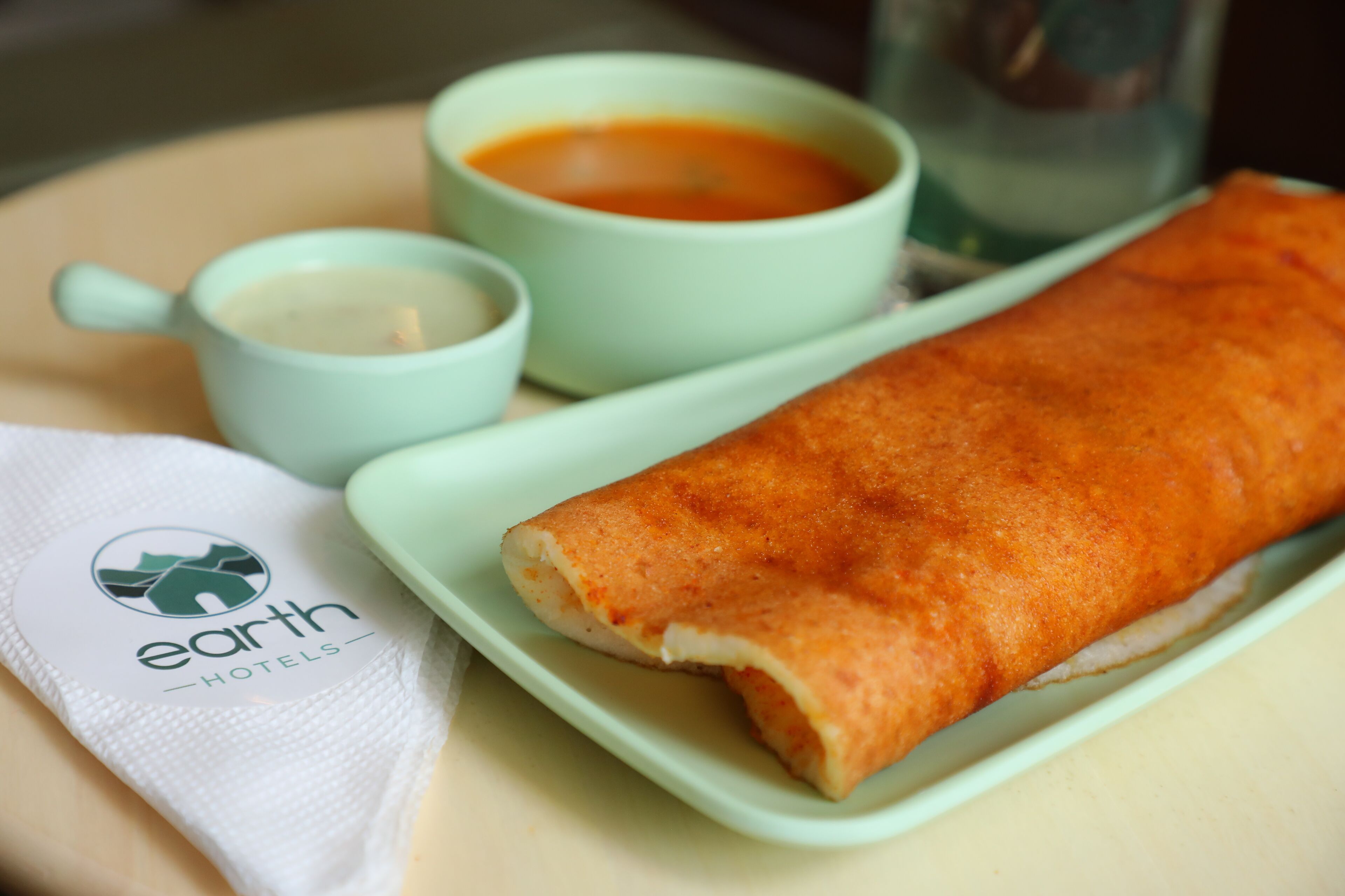 Une crêpe roulée servie sur une assiette verte, accompagnée d’une petite tasse de thé et d’une petite tasse de soupe, le tout disposé sur une table où se trouve une serviette ornée du logo de « earth hotels ».