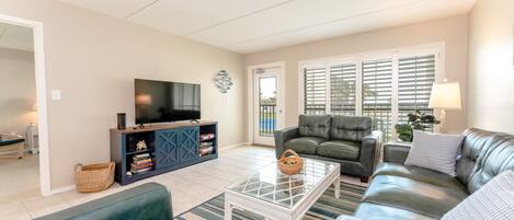 A living room with a sofa set, a glass coffee table, a TV on a cabinet, and a view through the window.