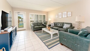 A living room with a TV, two green sofas, a coffee table, and a large window with blinds.