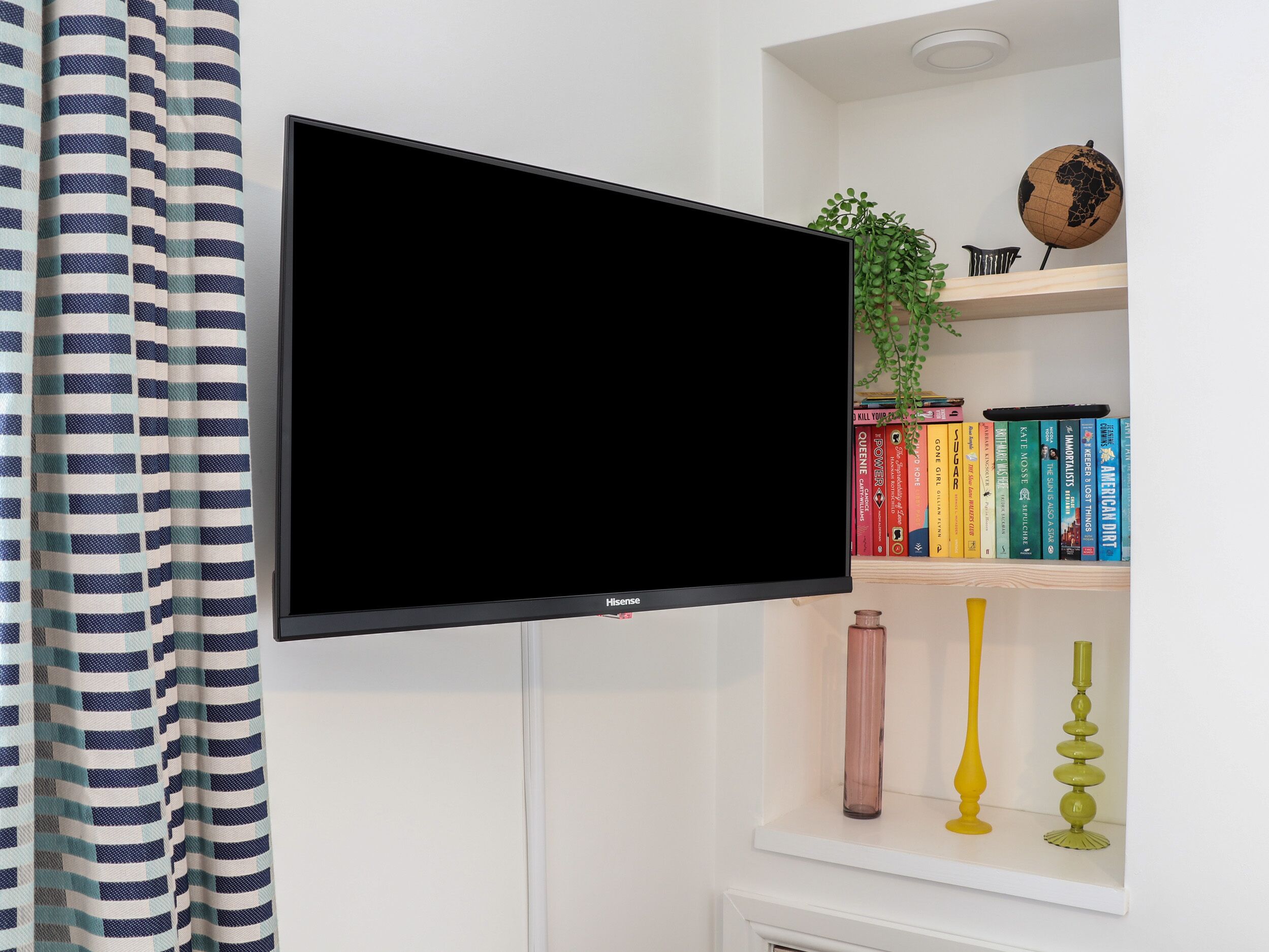 Un televisor de pantalla plana montado en una pared blanca, con una cortina a rayas a la izquierda y una estantería con libros y objetos decorativos a la derecha.