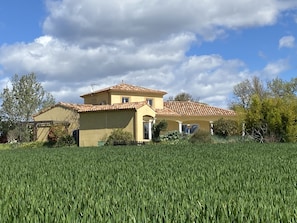 A two-story house with a tiled roof, surrounded by a green field and trees.