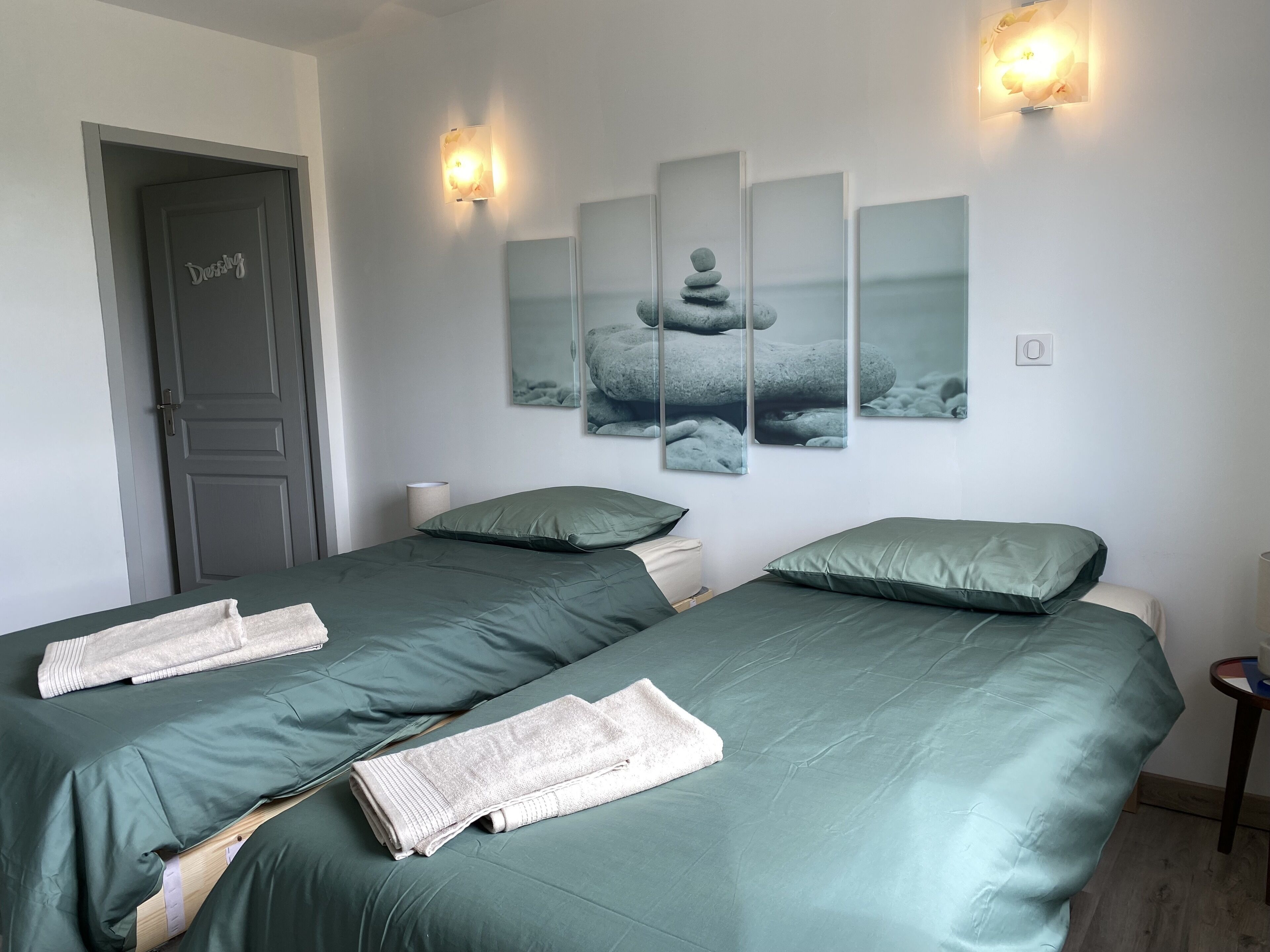 A room with two beds, each with green bedding and pillows, a door labeled "Dossing," and a wall-mounted artwork of stacked stones.