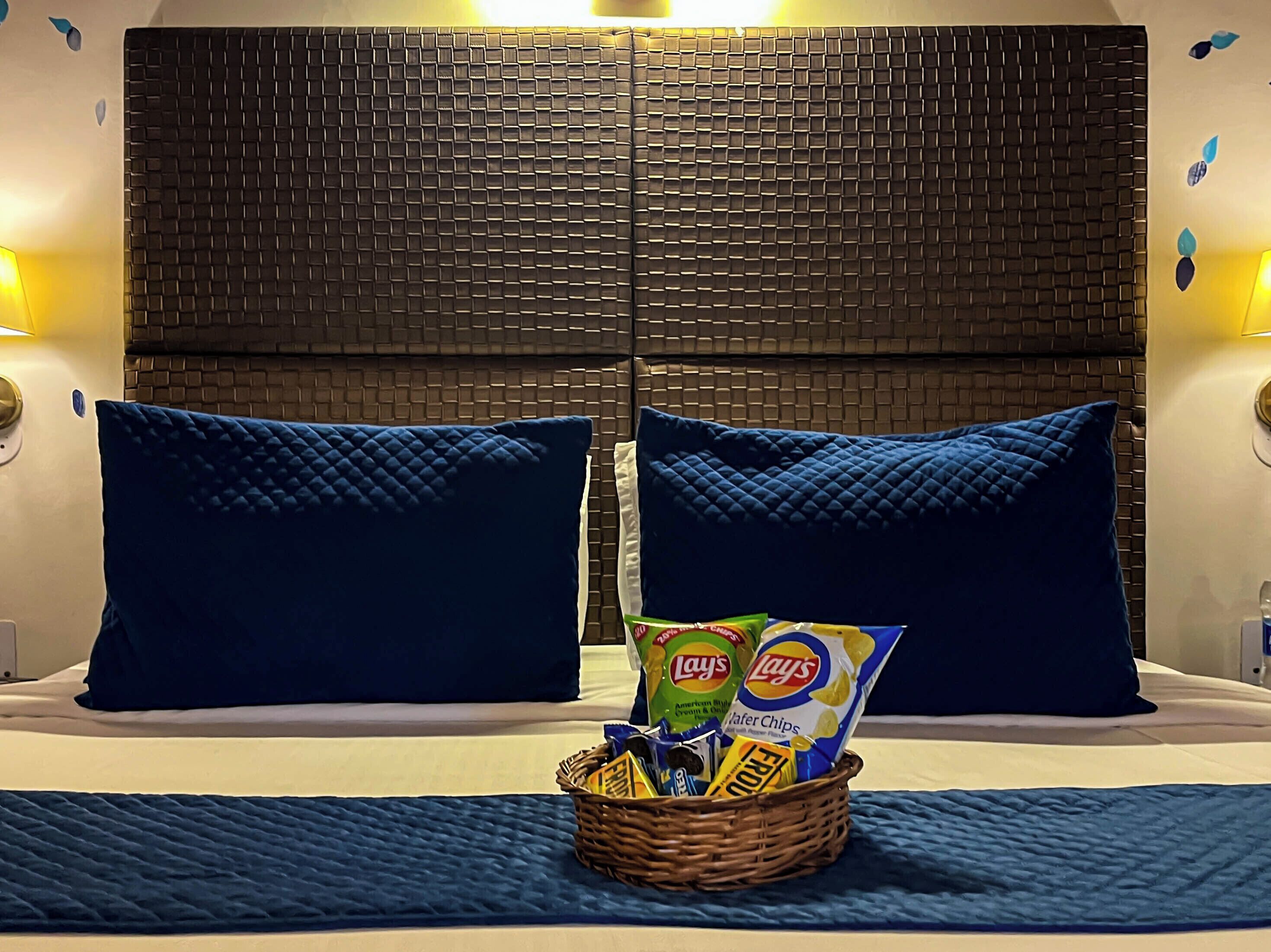 A hotel room with a bed, two blue pillows, a basket of snacks, and a textured wall behind the bed.