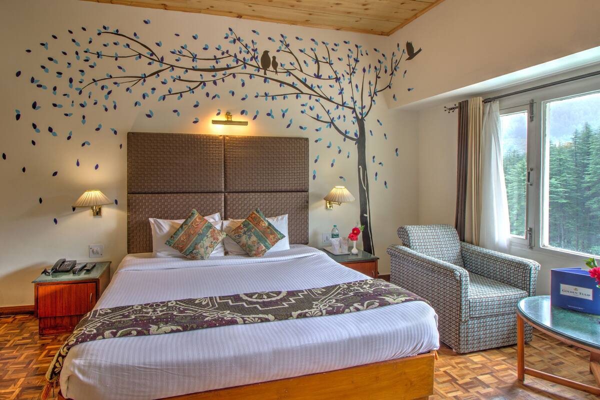 A hotel room with a large bed, a chair, a small table, a nightstand, and a window with a view of trees.