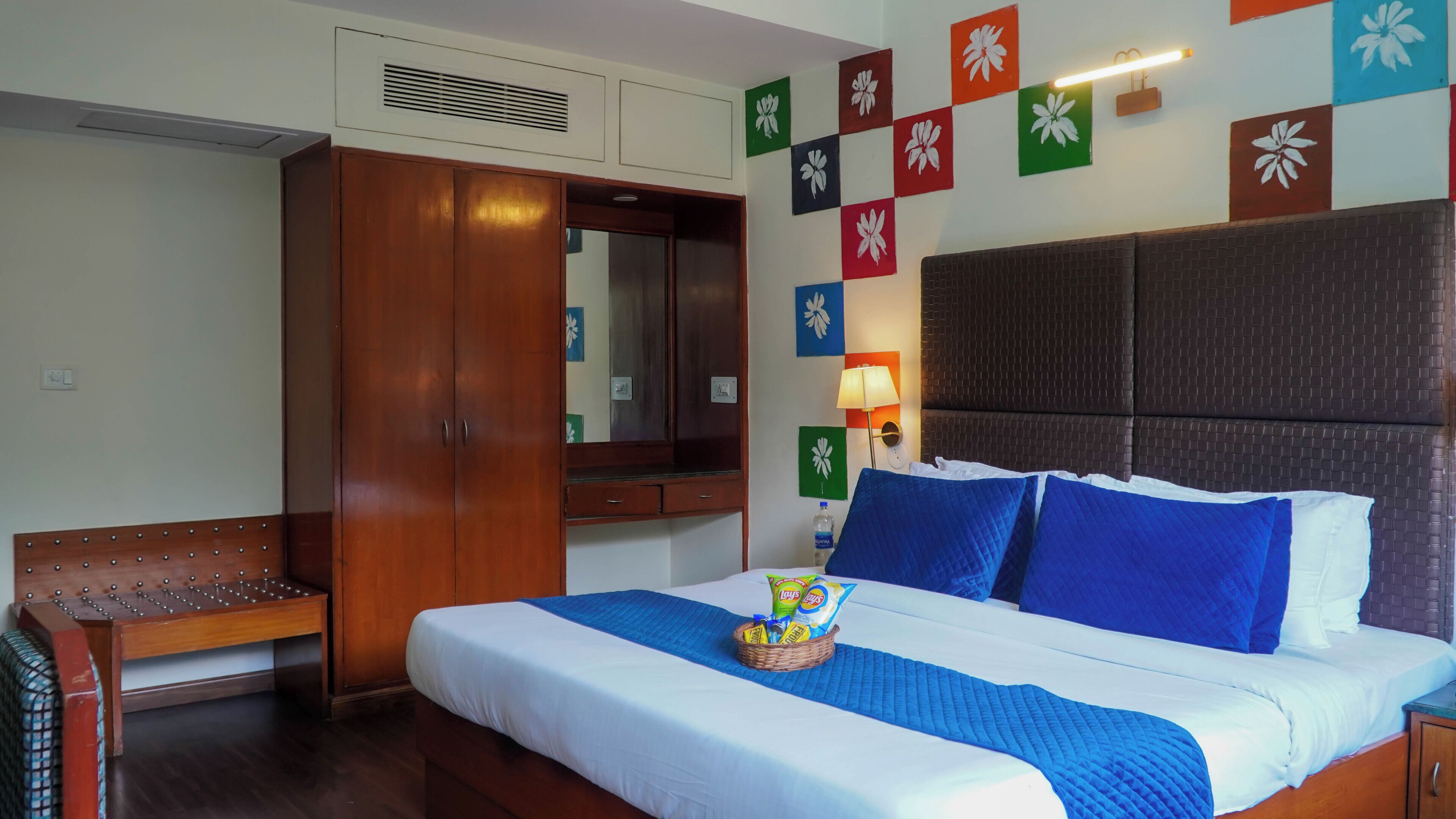 A hotel room with a bed, a wardrobe, a bench, and a wall with colorful floral decorations.