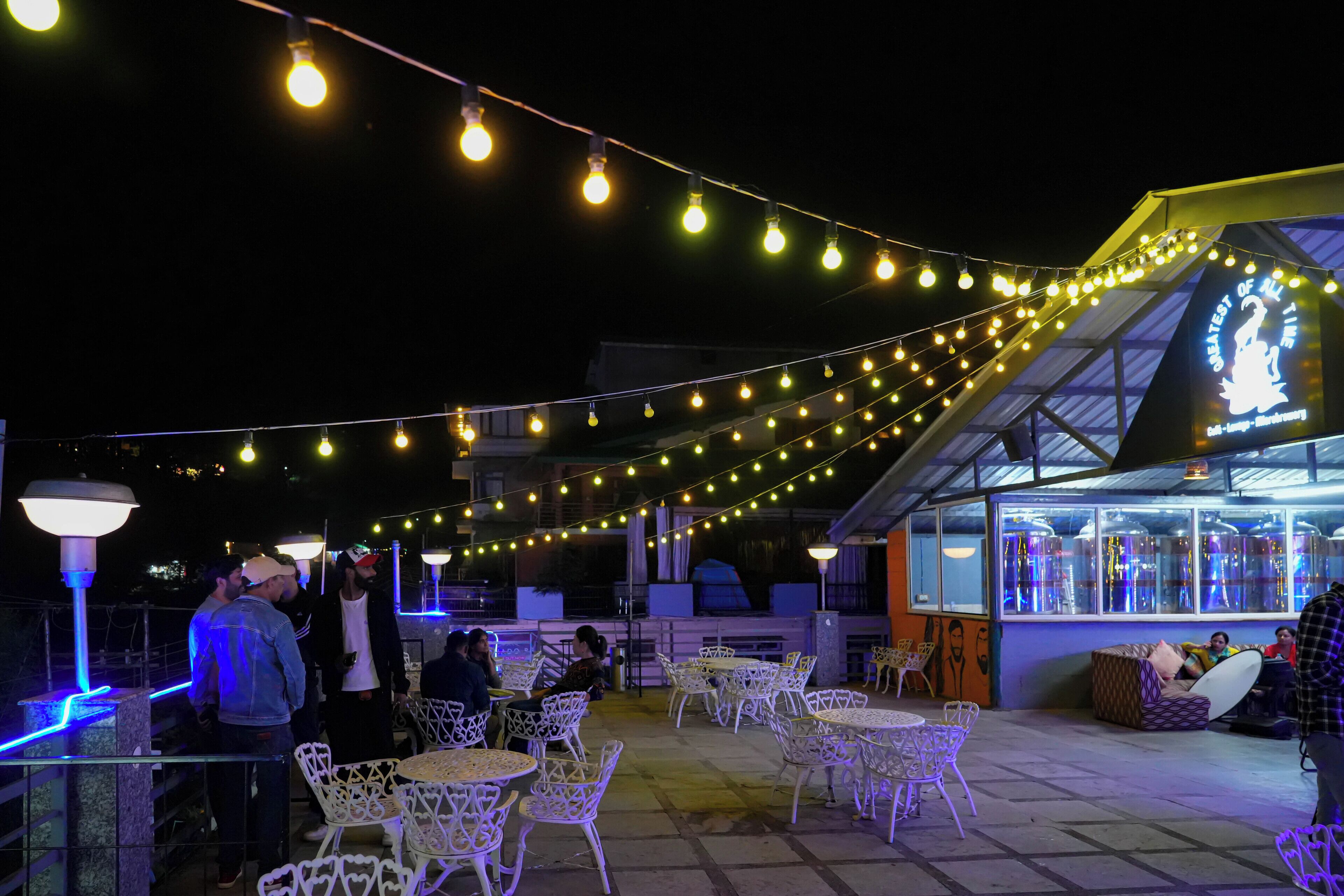 An outdoor seating area with tables and chairs, string lights overhead, and a building with a sign in the background.