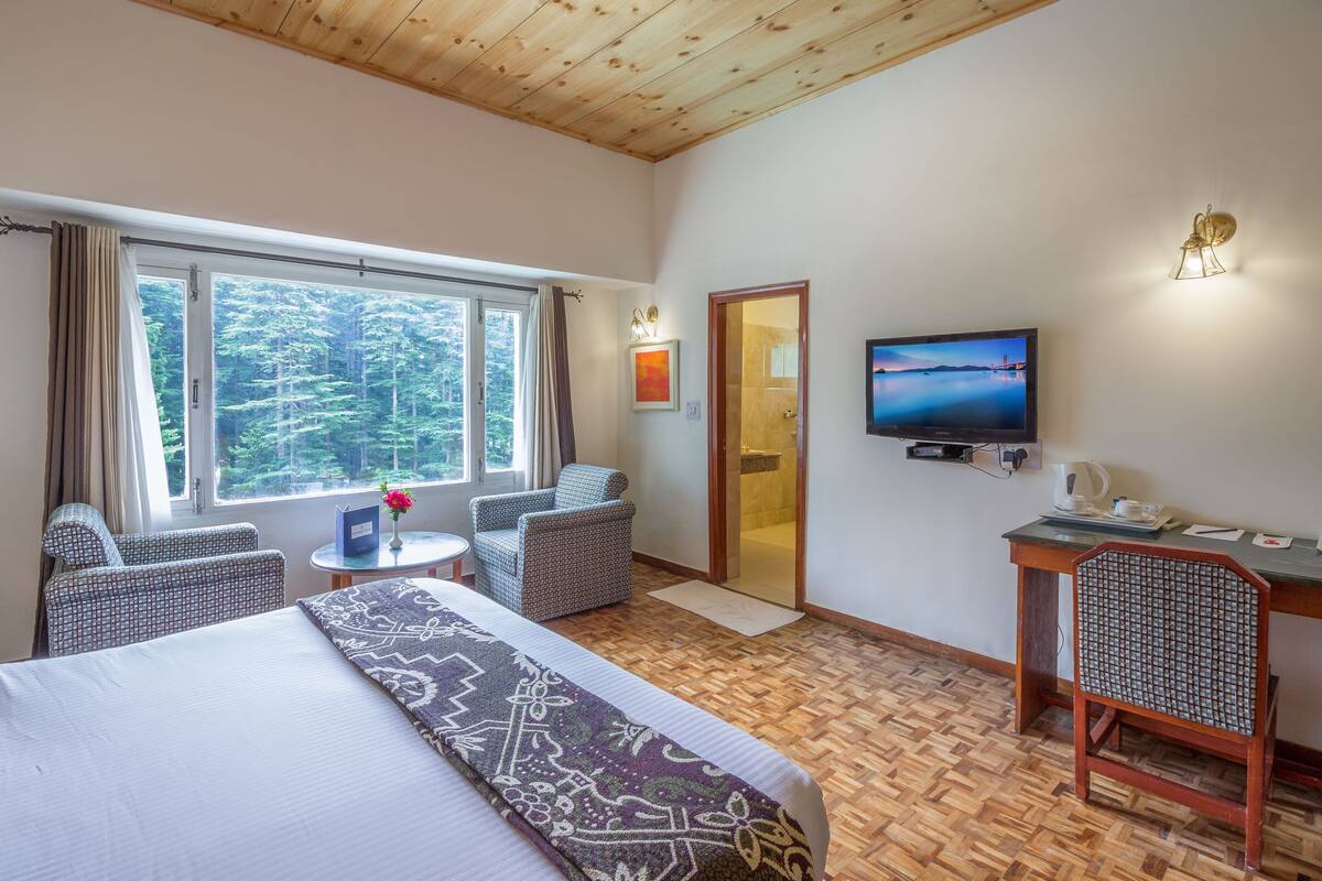 A hotel room with a bed, two patterned chairs, a small table with a vase, a television, and a view of a forest through a large window.