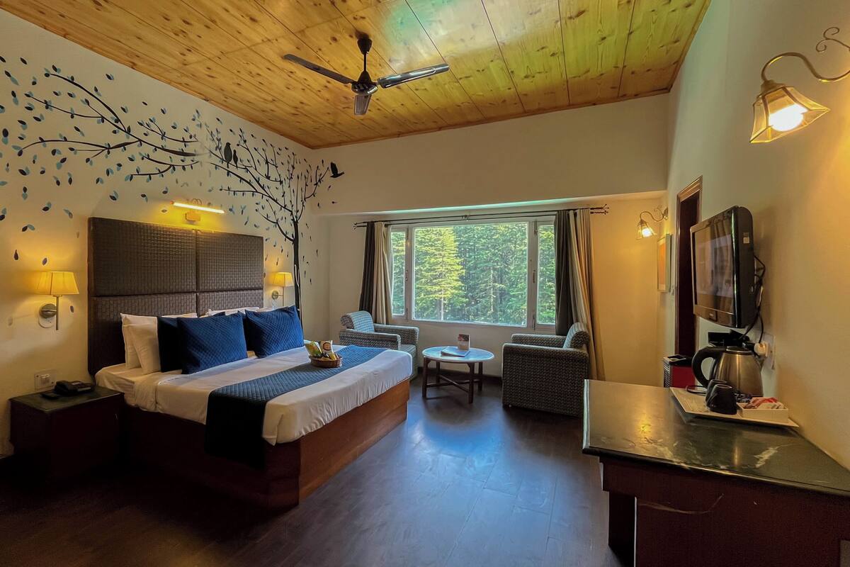 A bedroom with a bed, bedside lamps, a ceiling fan, a window with curtains, a small table, a chair, a television, and a coffee maker.