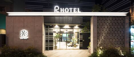 A hotel entrance with a modern facade, a visible logo, and a well-lit interior.