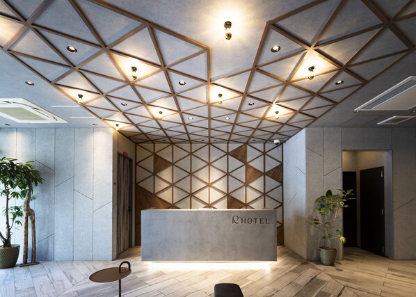 A hotel reception area with a geometric ceiling design, a reception desk labeled "RHOTEL," and potted plants.