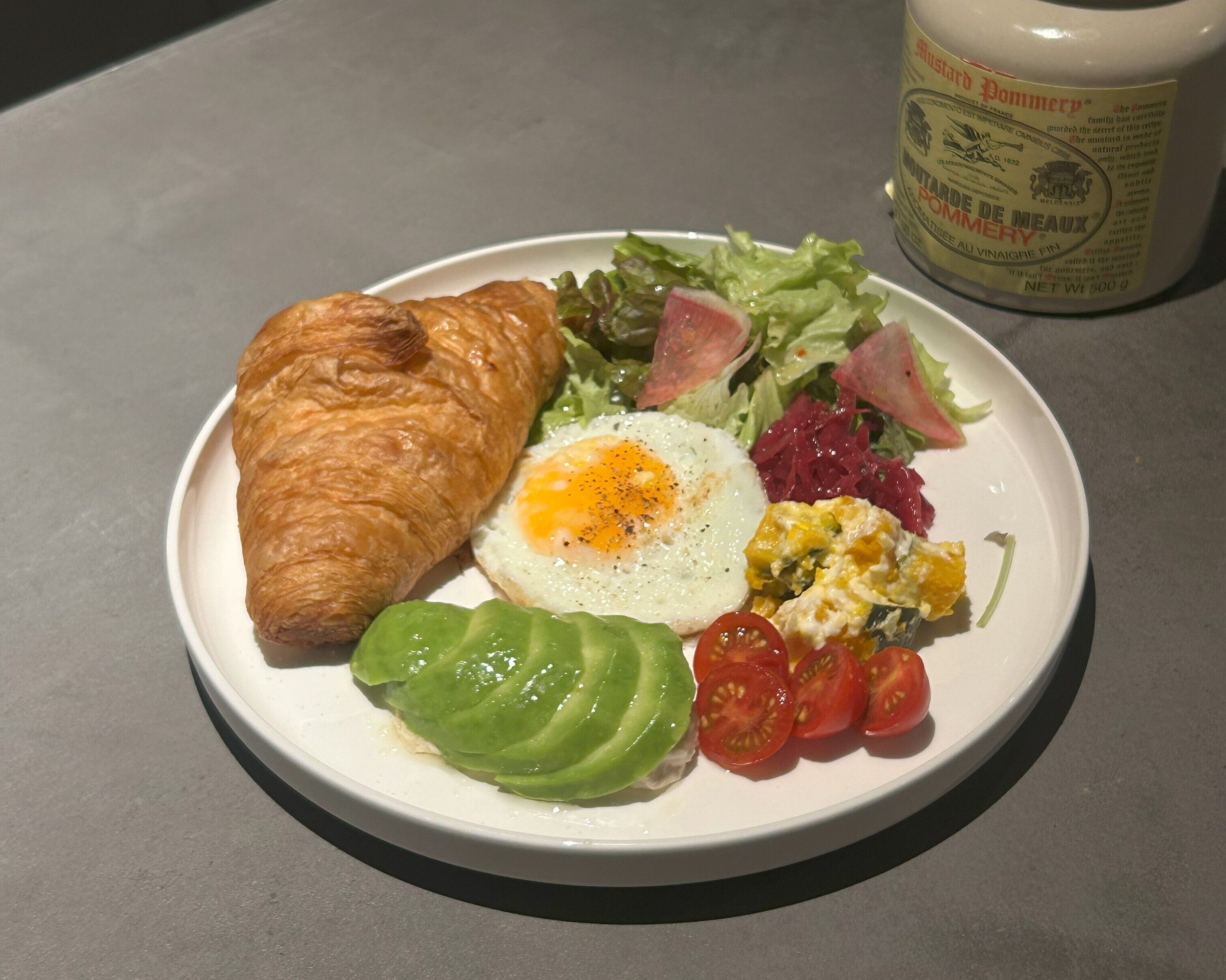 Ein Teller mit einem Croissant, einem Spiegelei, einem Salat und einer Senfpackung.
