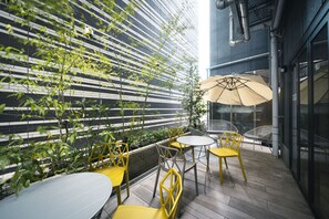 An outdoor patio with yellow and white tables and chairs, a beige umbrella, and a modern building with a striped facade in the background. - R Hotel Namba South (Osaka)