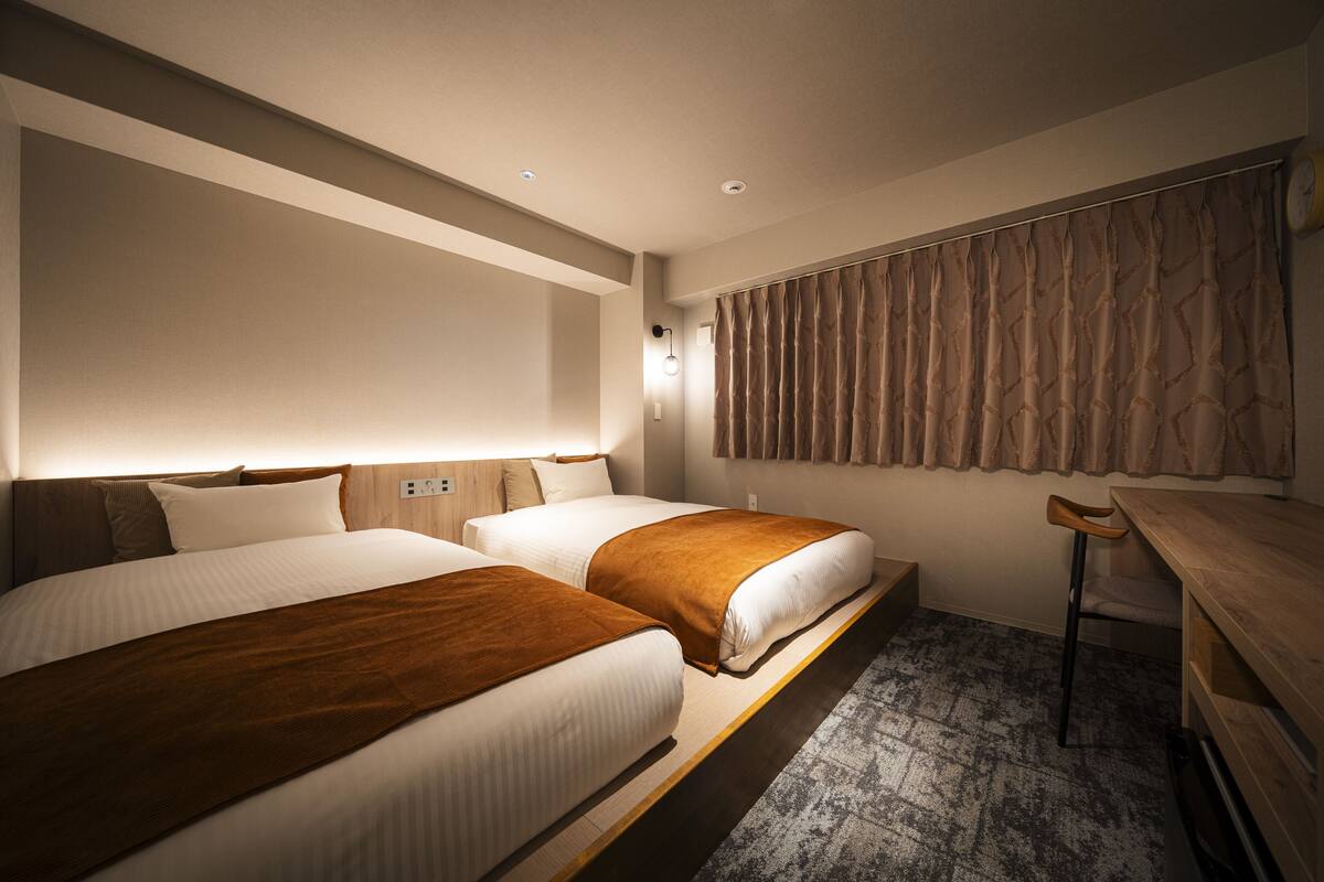 Habitación de hotel con dos camas, un escritorio de madera, una silla, un ventanal con cortinas y un reloj en la pared.