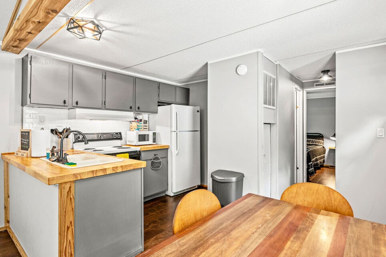 Een compacte keuken met grijze kastjes, een witte koelkast en een houten eettafel met stoelen.