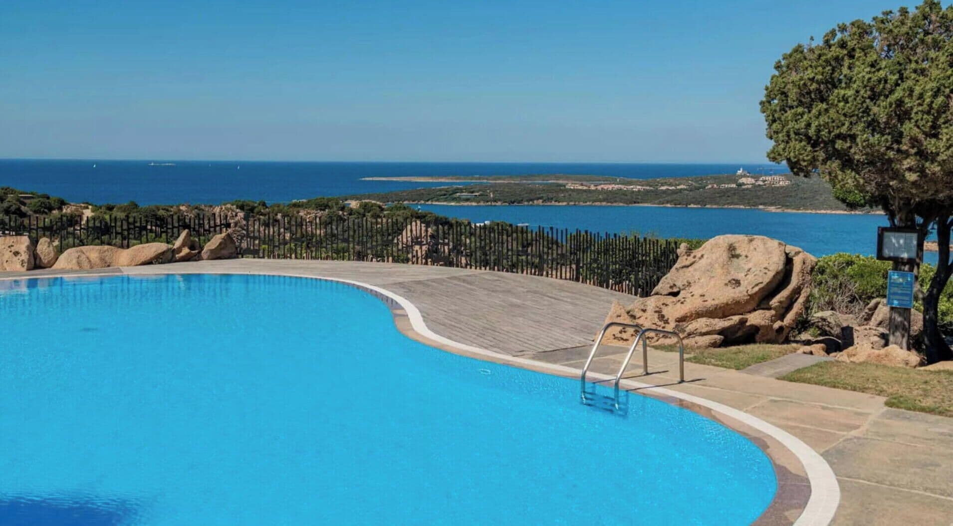 A clear blue swimming pool with a wooden deck, surrounded by a fence and large rocks.