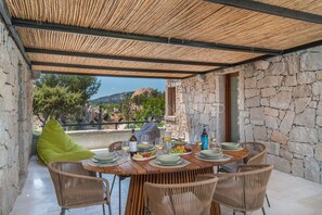 An outdoor dining area with stone walls, a thatched roof, wicker chairs, and a view of the landscape. - Villetta Ashlar - SARDINIA UNLIMITED (Porto Cervo)