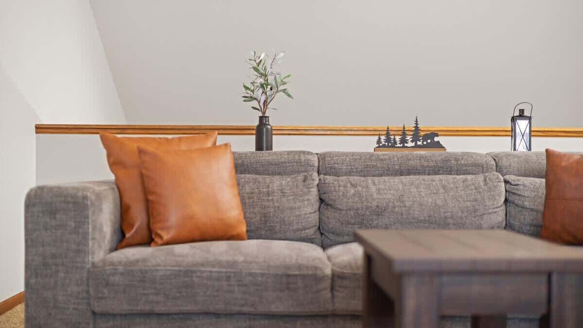 A grey sectional sofa with two brown cushions, a small table, a vase with green plants, and a decorative wall piece.