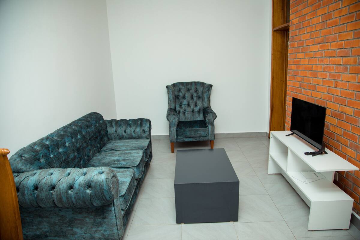 A living room with a teal sofa, a matching armchair, a black coffee table, and a TV on a white stand against a brick wall.