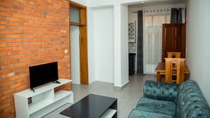 A living room with a brick wall, a flat-screen TV on a white stand, a blue sofa, and a wooden dining table with chairs. -  JLD Apartments (Kigali)