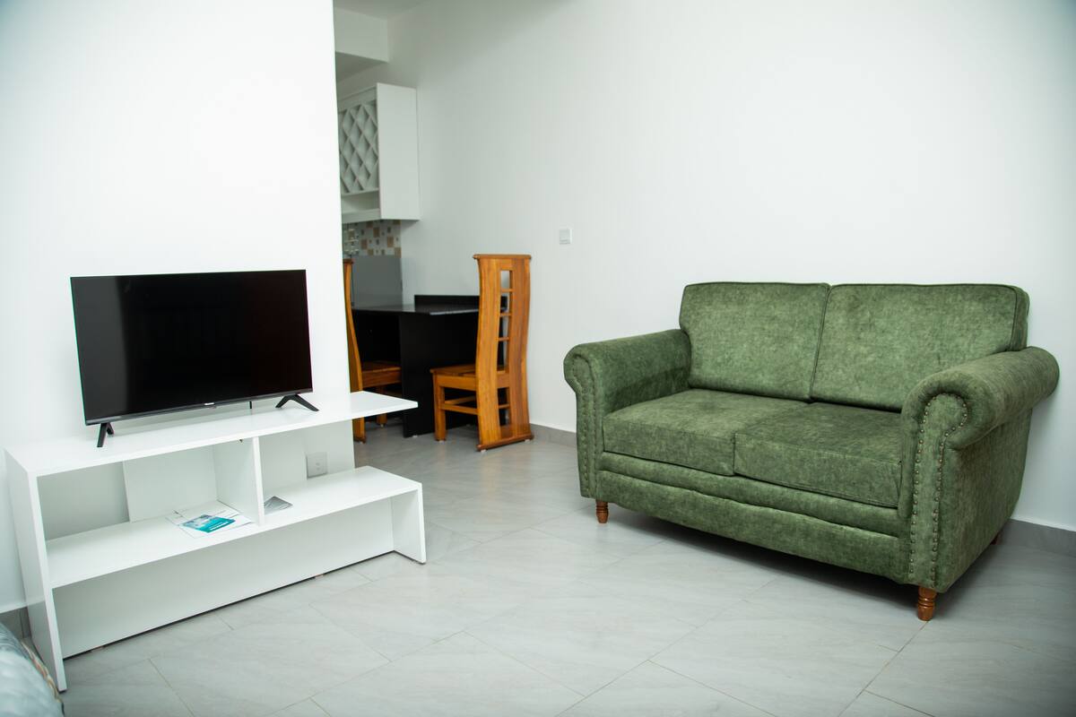 A living room with a flat-screen TV on a white stand, a green sofa, and a wooden chair.