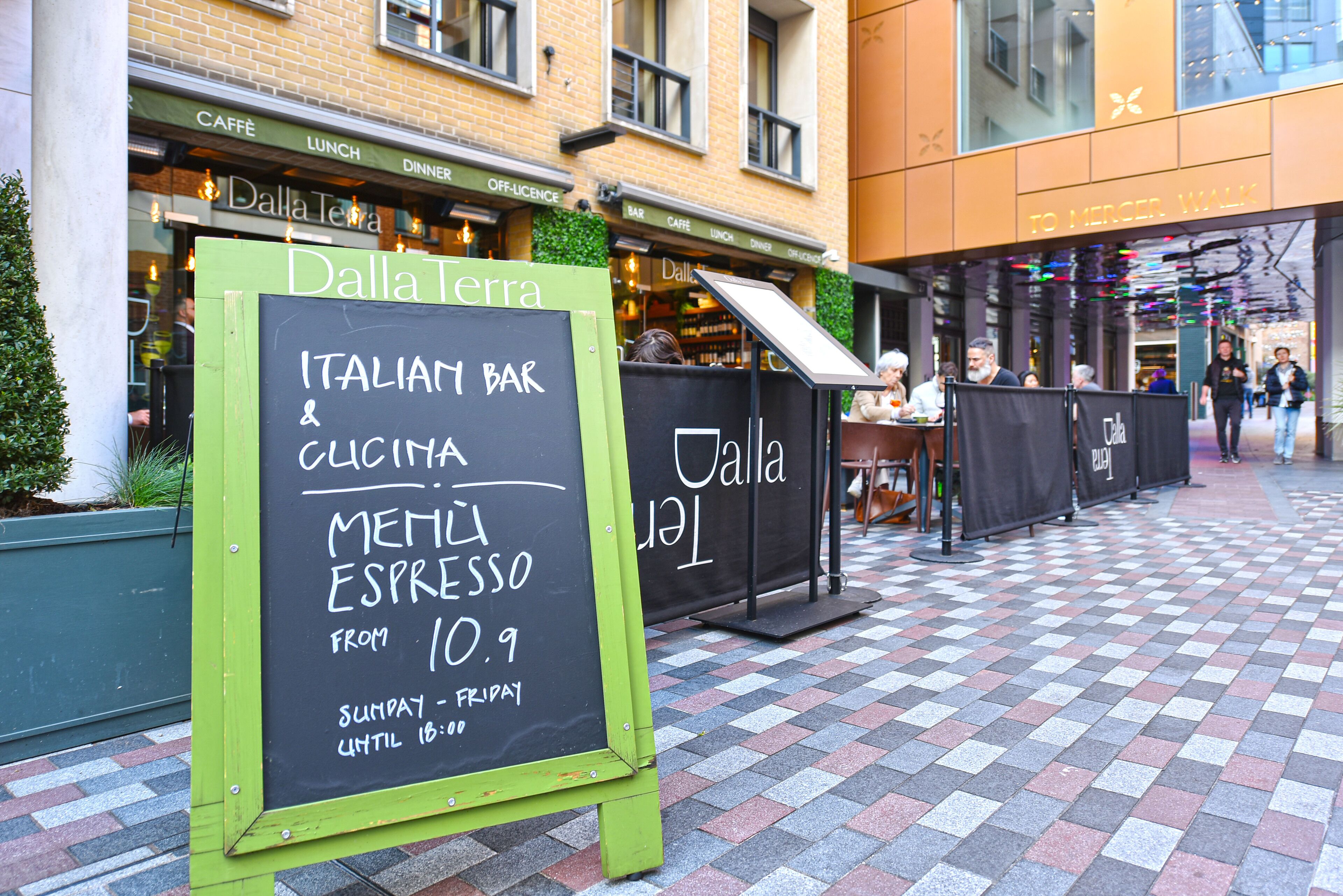Un espace extérieur aménagé avec des sièges et un tableau noir servant d’enseigne pour un bar italien, où sont également indiqués les plats proposés.
