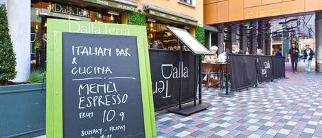 Un espace extérieur aménagé avec des sièges et un tableau noir servant d’enseigne pour un bar italien, où sont également indiqués les plats proposés.