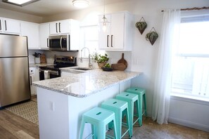 Una cocina moderna con armarios blancos, electrodomésticos de acero inoxidable, encimera de granito y taburetes de bar color aguamarina.