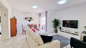 Uma sala de estar moderna com um sofá branco, uma mesa de jantar com cadeiras, uma televisão de tela plana e um vaso com planta.