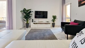 Uma sala de estar moderna com um sofá branco, um sofá preto, uma televisão de tela plana, um rack branco e duas plantas em vasos.