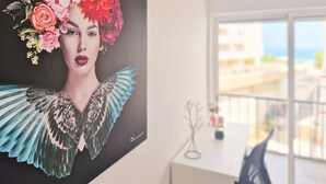 Quarto com uma grande obra de arte floral na parede, uma escrivaninha branca e vista para o mar através de grandes janelas.
